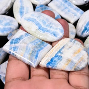 Puede incluir: Una colección de piedras preciosas pulidas en varias formas, incluyendo ovaladas, rectangulares e irregulares. Las piedras presentan un llamativo patrón de vetas azules y blancas, con toques de amarillo y beige.