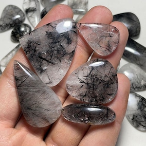 May include: A collection of polished gemstones, held in a hand, featuring clear quartz with black tourmaline inclusions. The stones vary in shape, including teardrop, oval, and triangular forms. The background shows additional gemstones.