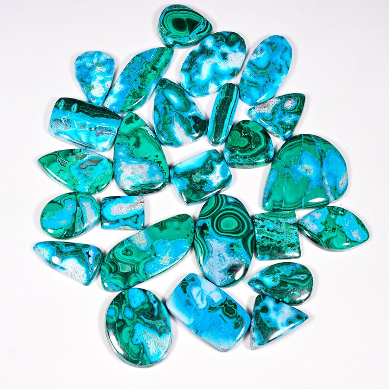 May include: A collection of polished gemstones in various shapes, including ovals, teardrops, and rectangles. The stones display a mix of vibrant turquoise and deep green hues, with some featuring intricate patterns and swirls. The gemstones are arranged on a white surface.