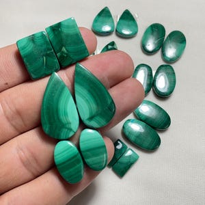 May include: A collection of polished malachite gemstones in various shapes, including teardrops, ovals, and squares. The stones display a vibrant green color with distinctive banded patterns. The gemstones are displayed on a light-colored surface.