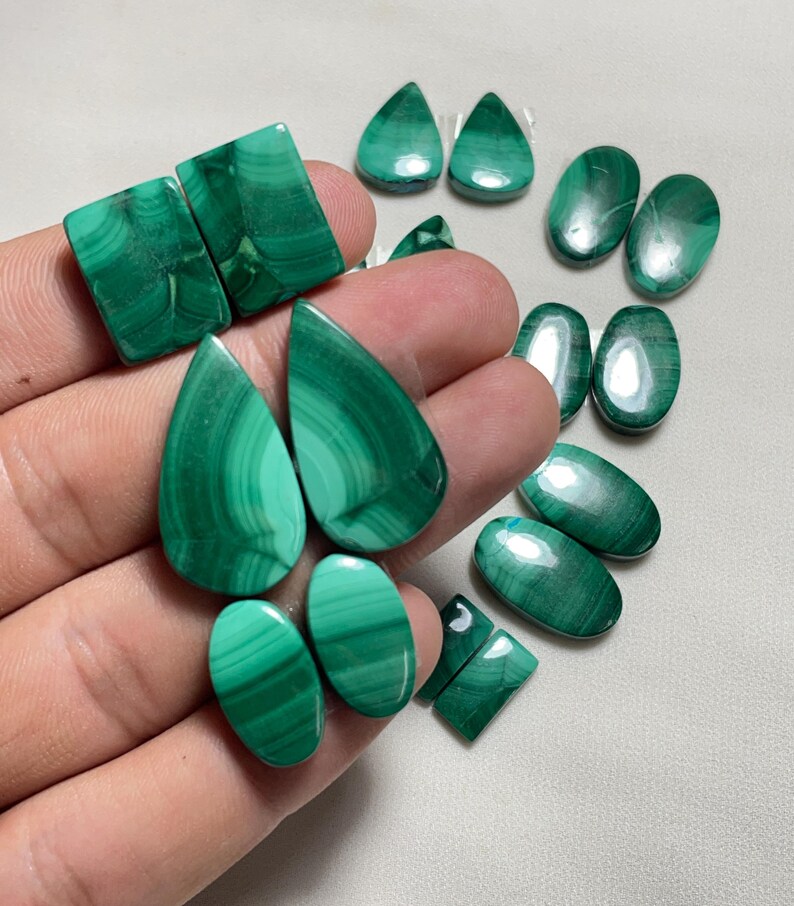May include: A collection of polished malachite gemstones in various shapes, including teardrops, ovals, and squares. The stones display a vibrant green color with distinctive banded patterns. The gemstones are displayed on a light-colored surface.