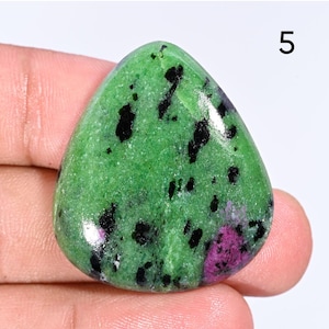 May include: A polished, teardrop-shaped gemstone with a vibrant green base, speckled with black inclusions and a patch of deep pink. The number "5" is visible in the upper right corner. This natural stone is likely a type of Zoisite with Ruby.