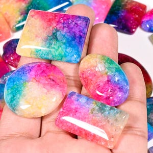 May include: A selection of polished gemstones in diverse shapes, including round, oval, and rectangular. The stones exhibit a vibrant array of colours, including pink, yellow, blue, and purple. The gemstones are held in a hand, highlighting their smooth, glossy surfaces.