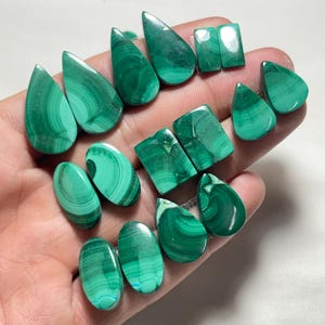 May include: A collection of polished malachite gemstones in various shapes, including teardrops, ovals, and squares. The gemstones display a vibrant green color with unique swirling patterns. The stones are held in a hand, showcasing their natural beauty and variations.