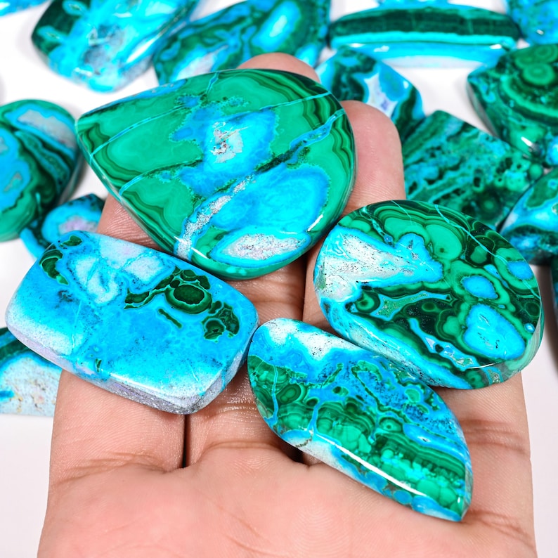 May include: A collection of polished gemstones with a striking mix of green and blue hues. The stones display intricate patterns and textures, with a polished, glossy finish. The gemstones are held in a hand, showcasing their natural beauty.