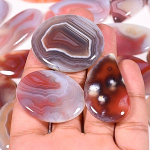 Puede incluir: Una colección de piedras de ágata pulidas de varias formas y colores. Las piedras presentan bandas de color blanco, rojo, marrón y gris, con patrones únicos. Una piedra tiene forma de lágrima, otras son ovaladas y redondas.