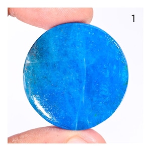 Pode incluir: Uma pedra preciosa redonda e polida com uma cor azul vibrante e sólida. A pedra tem uma superfície lisa e é segurada por um dedo. O número "1" é visível no canto superior direito. Esta pedra preciosa é provavelmente usada para joias ou fins decorativos.