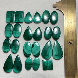May include: A collection of polished malachite gemstones in various shapes, including ovals, teardrops, and rectangles. The stones display rich green hues with distinctive swirling patterns. A brass ruler is positioned to the right, providing scale.