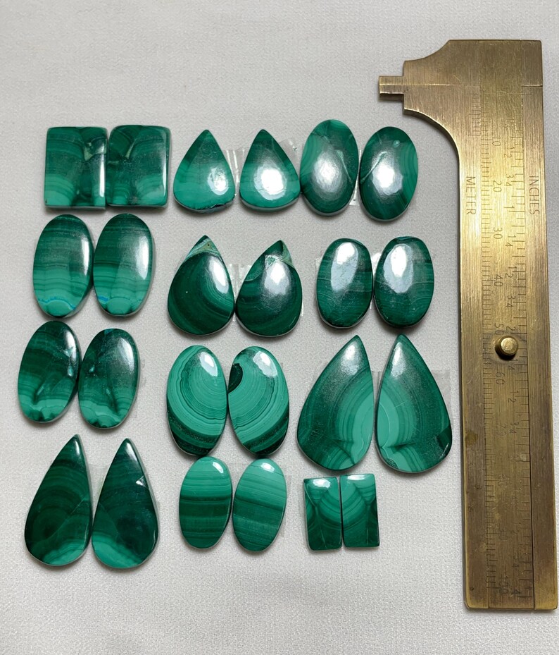 May include: A collection of polished malachite gemstones in various shapes, including ovals, teardrops, and rectangles. The stones display rich green hues with distinctive swirling patterns. A brass ruler is positioned to the right, providing scale.