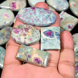 May include: A collection of polished gemstones in various shapes, including oval and rectangular. The stones feature a light blue-green matrix with vibrant pink and red inclusions. The gemstones are held in a hand.