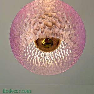 Può includere: Una lampada a sospensione sferica rosa con un motivo geometrico strutturato. La luce ha un bordo intrecciato color oro attorno all'apertura. La lampada è appesa al soffitto. L'indirizzo del sito web ilodecor.com è visibile in basso.
