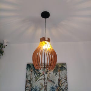 Może przedstawiać: Drewniana lampa wisząca z włączoną żarówką. Lampa ma kształt łzy i jest wykonana z naturalnego drewna. Lampa jest zawieszona na czarnym przewodzie.