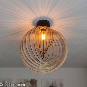 Op de afbeelding: Een houten, bolvormige plafondlamp met een warme, gloeiende gloeilamp in het midden. Het licht wordt omgeven door concentrische, gebogen houten stroken, waardoor een uniek, open ontwerp ontstaat. De lamp is gemonteerd op een donkerbruine basis.
