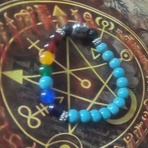 May include: A beaded bracelet featuring a chakra design. The bracelet includes a variety of colorful beads, including red, orange, yellow, green, blue, and purple. The bracelet also has turquoise and black beads. The bracelet is displayed on a brown surface with a gold design.