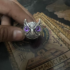 Könnte beinhalten: Ein silberner Eulenring mit violetten Augen. Der Ring befindet sich an einem Finger und der Hintergrund ist eine Tarotkarte mit dem Text "The Magician".