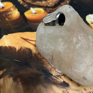 Puede incluir: Un anillo de plata con una piedra negra engastada sobre una superficie clara y texturizada. El anillo tiene un diseño simple y clásico. El fondo incluye una imagen borrosa de velas y otros objetos.