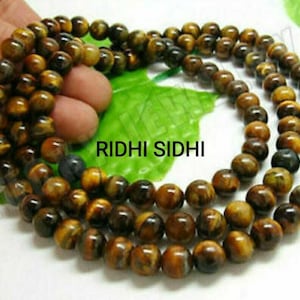 Może przedstawiać: Długi sznur brązowych i złotych koralików z tygrysiego oka. Koraliki są okrągłe i mają gładkie, polerowane wykończenie. Sznur składa się z wielu rzędów koralików, tworząc warstwowy efekt. Koraliki są nawleczone na przezroczysty sznurek.