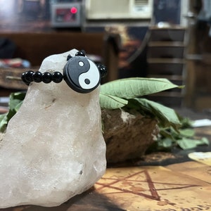 May include: A black beaded bracelet with a white and black yin yang charm. The bracelet is resting on a white rock.