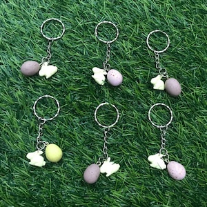 May include: Six keyrings with silver rings and chains. Each keyring has a small white bunny charm and a small egg charm. The eggs are different colours, including purple, pink, and yellow.