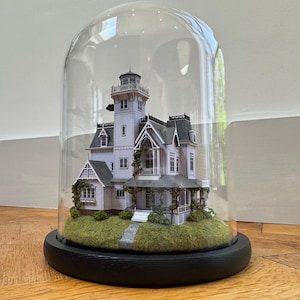 May include: A miniature Victorian-style house model, enclosed in a glass dome. The house is painted light gray with dark gray trim, a tower, and a porch. The base is black, and the house sits on a bed of green moss. The dome is clear.
