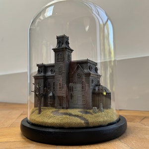 May include: A miniature model of a spooky Victorian house with a glass conservatory, set in a diorama under a glass dome on a black wooden base.
