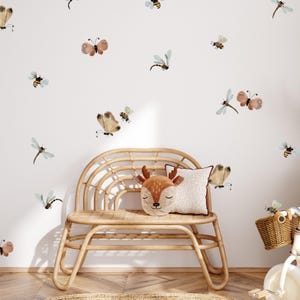 Può includere: Un muro bianco con una varietà di adesivi murali di farfalle e libellule colorati. Una panca in vimini marrone chiaro con un cuscino a forma di cervo marrone si trova di fronte al muro.