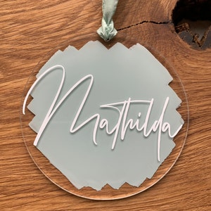 Könnte beinhalten: Ein runder, transparenter Acryl-Anhänger mit einem hellblauen, bemalten Hintergrund und dem Namen "Mathilda" in weißer Schrift.