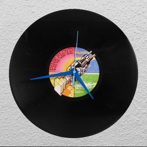 Puede incluir: Un reloj de vinilo negro con manecillas azules, con la carátula del álbum "Wish You Were Here" de Pink Floyd. La etiqueta colorida muestra un diseño surrealista. El reloj está montado en una pared blanca.