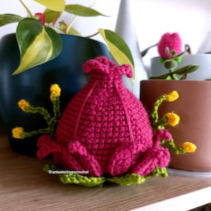 Puede incluir: Una flor rosa de crochet con detalles amarillos y verdes. La flor está sentada en un estante de madera con otras plantas en el fondo.
