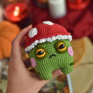 May include: A small, green, crocheted creature with a red mushroom cap featuring white spots. The figure has large, dark eyes with yellow borders, a small embroidered mouth, and light pink circular patches on its cheeks. It is a soft, stuffed toy.