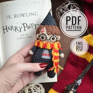 以下が含まれることがあります： かぎ針編みのハリーポッター人形。傷跡、メガネ、グリフィンドールのスカーフ付き。人形は黒いローブと尖った帽子を着用しています。画像には「ハリー・ポッターと炎のゴブレット」というタイトルの本と杖が含まれています。