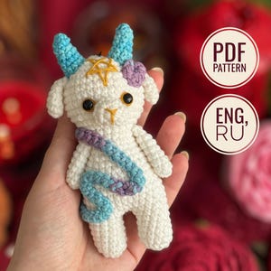 May include: A white crocheted goat doll with blue horns, a yellow pentagram embroidered on its forehead, and a purple and blue crocheted snake draped across its body. The doll has black eyes and includes text indicating a PDF pattern in English and Russian.