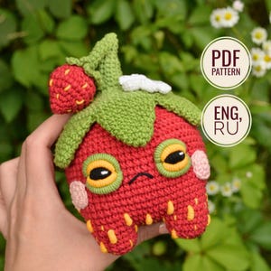 Puede incluir: Una criatura de fresa tejida a ganchillo en rojo con una parte superior de hojas verdes, un pequeño detalle de fresa y semillas amarillas. Tiene ojos grandes con bordes amarillos, mejillas rosadas y un ceño fruncido. Texto: PDF PATTERN ENG, RU.