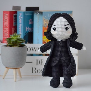 May include: A handmade crochet doll of a character with black hair and a black coat. The doll has a white face with black eyes and is standing in front of a white wooden crate with books and a small succulent plant in a gray pot.