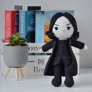 May include: A handcrafted crochet doll featuring a white face, black hair, and a black ensemble. The doll is positioned before a white wooden crate with books and a small potted succulent. The doll's attire includes a long coat and buttoned shirt.