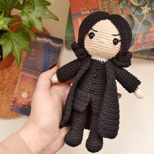 May include: A handmade crochet doll of a character with black hair and a black coat. The doll has a neutral skin tone, black eyes, and a white collar. The doll is holding a black coat and black trousers.