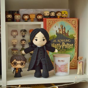 May include: A shelf arrangement featuring Harry Potter merchandise. A large, crocheted Professor Snape doll is the focal point, with a Harry Potter Funko Pop figure nearby. A hardback book, a candle, and other figurines are also present.
