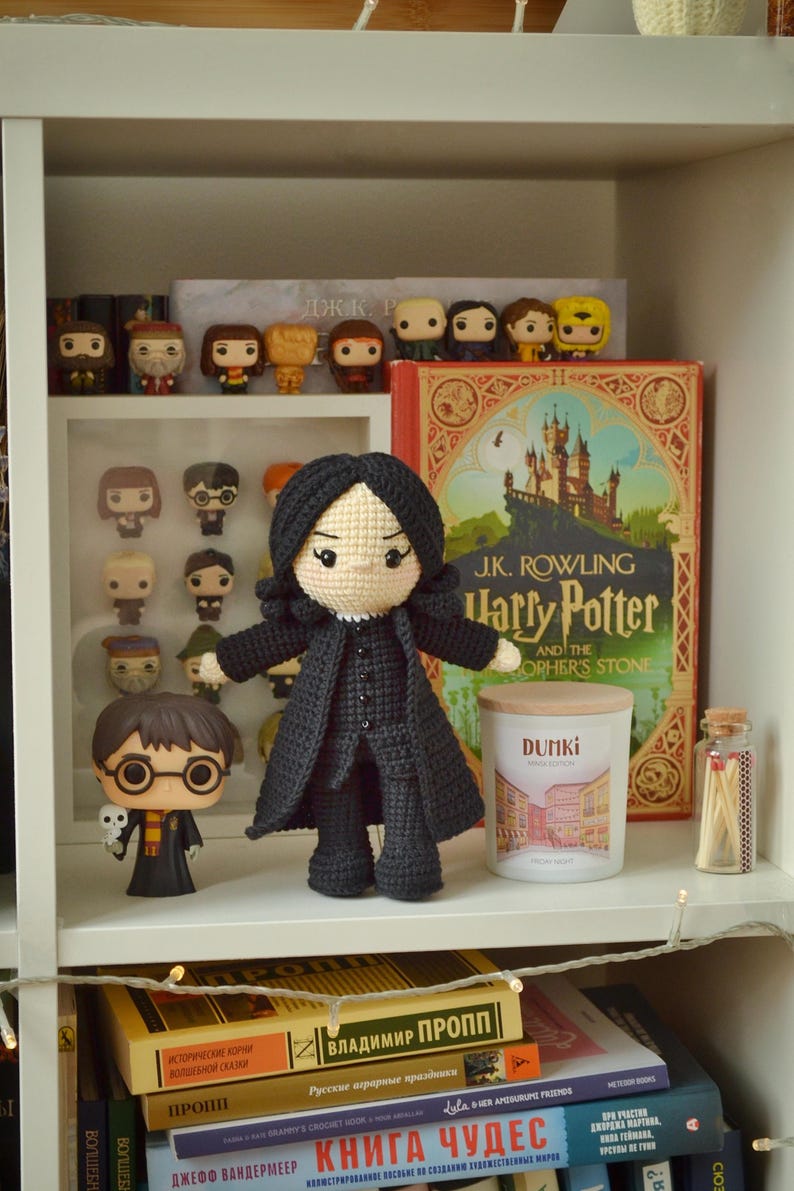 May include: A shelf arrangement featuring Harry Potter merchandise. A large, crocheted Professor Snape doll is the focal point, with a Harry Potter Funko Pop figure nearby. A hardback book, a candle, and other figurines are also present.