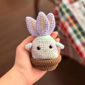 Puede incluir: Una planta de ganchillo hecha a mano con una cara sonriente, ojos negros y brazos pequeños. La planta tiene una cabeza de color púrpura claro con detalles bordados en amarillo y una maceta marrón. La planta mide aproximadamente 10 cm de altura.