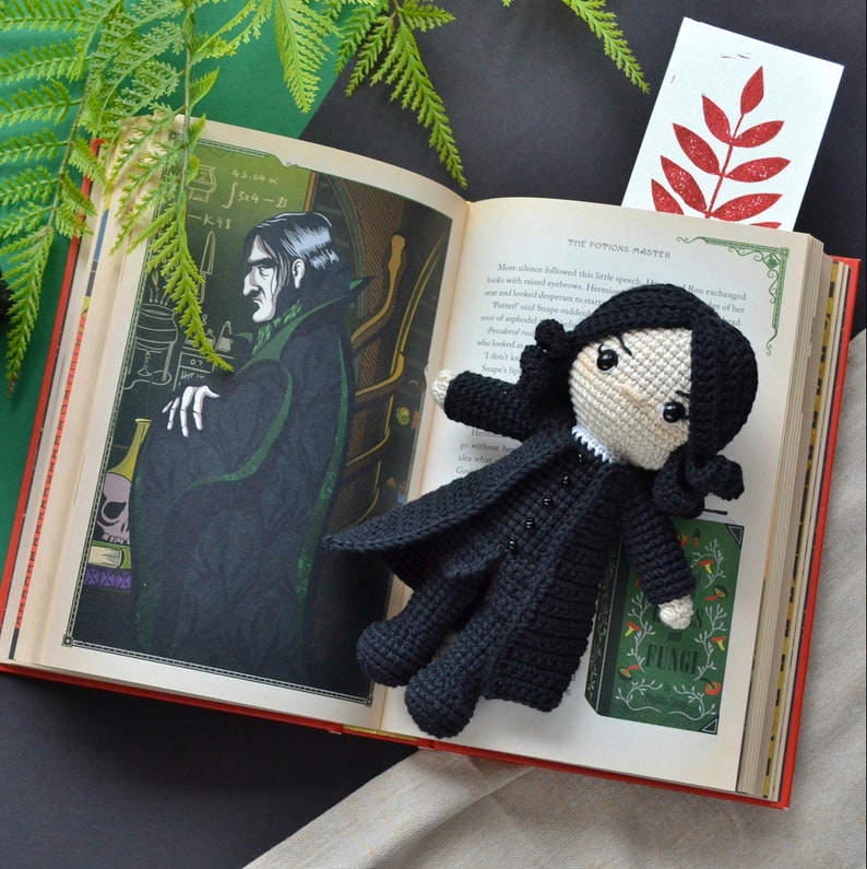 May include: A black crochet doll with a long coat and white collar, lying on an open book with illustrations. The doll is placed on a book page with text and a green and black background.