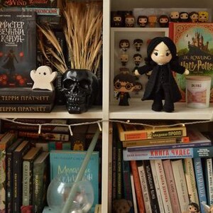 May include: A white bookcase filled with decorative items. A black skull vase contains dried wheat. A white ghost figure sits on books. A crocheted doll in a black coat stands near Harry Potter books and figurines.