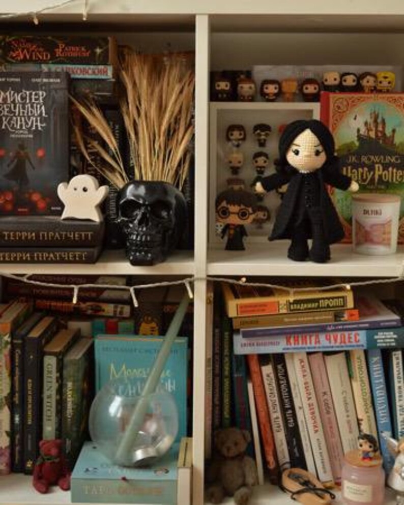 May include: A white bookcase filled with decorative items. A black skull vase contains dried wheat. A white ghost figure sits on books. A crocheted doll in a black coat stands near Harry Potter books and figurines.