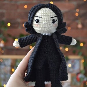 May include: A handmade crochet doll of a character with black hair and a black coat. The doll has a white face with black eyes and a stern expression. The doll's arms and hands are white. The doll is approximately 15 cm tall.