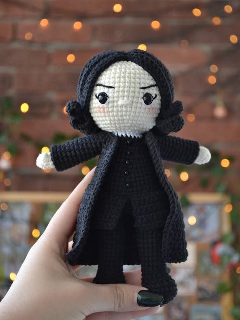 May include: A handmade crochet doll of a character with black hair and a black coat. The doll has a white face with black eyes and a stern expression. The doll's arms and hands are white. The doll is approximately 15 cm tall.