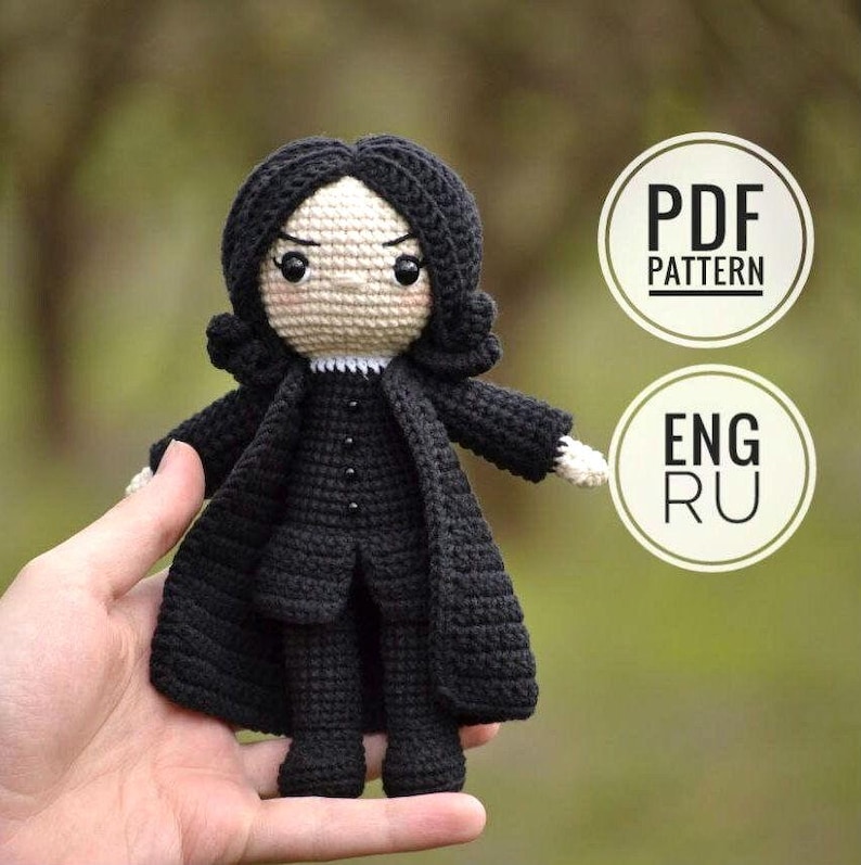 A hand holding a small crocheted amigurumi doll of a male professor character in a dark black robe. It features stylized black hair, pale skin, and a detailed frown. The doll stands against a blurred natural green background. Small icons indicate a PDF pattern in English and Russian.