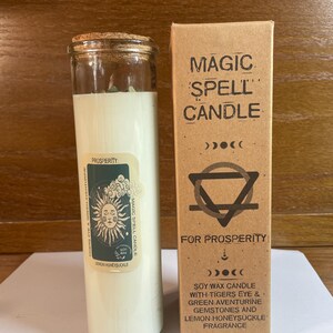 May include: A tall cylindrical glass candle with a cork lid and a cream-colored wax interior. The label features a sun illustration and the text "PROSPERITY". Beside it stands a brown cardboard box labeled "MAGIC SPELL CANDLE FOR PROSPERITY".
