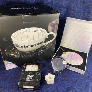 Könnte beinhalten: Ein Wahrsage-Teeservice mit einer weißen Teetasse und Untertasse, mit Symbolen und dem Text "Telling fortunes in tea". Enthält eine Kristallkugel, ein Pendel, Kerzen und einen sternförmigen Wachswärmer. Auf der Schachtel steht "Magic Spell Candles Happiness."