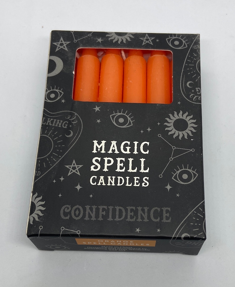 May include: A box of orange magic spell candles for confidence. The box has a black background with white stars, a sun, and an eye design. The text on the box reads "Magic Spell Candles" and "Confidence".