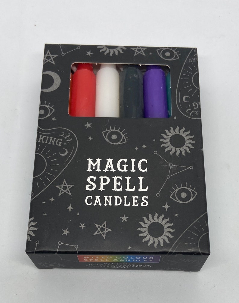 May include: A box of Magic Spell Candles with a black background and silver stars, moons, and eyes. The box contains six candles in red, white, black, purple, and teal.