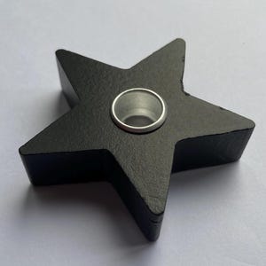 May include: A matte black star-shaped candle holder with a smooth finish and a circular silver-toned metal insert in the center. The object features five distinct points and a flat top surface, designed to hold a single small candle securely.